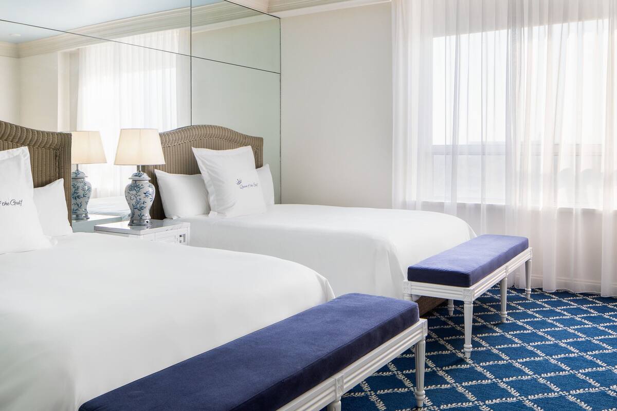 A hotel room with two beds, a bench, a nightstand with lamps, and a large mirror.