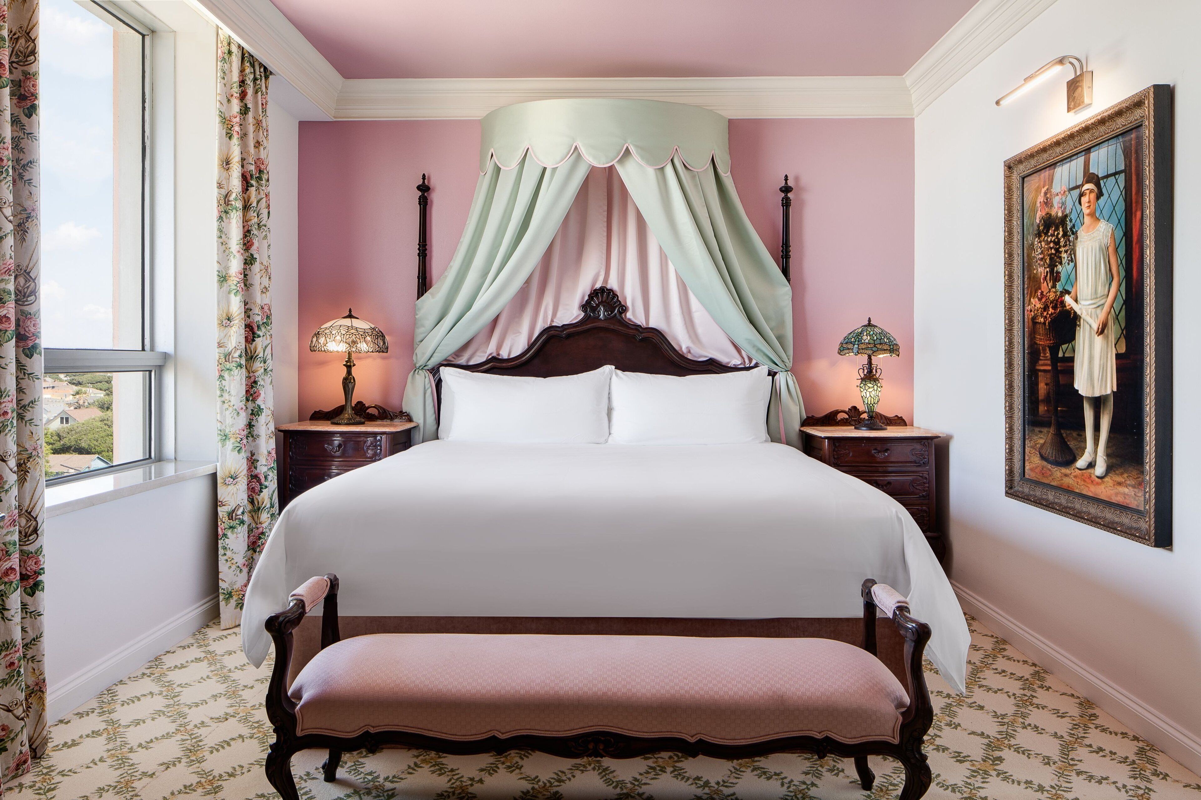 A bedroom with a four-poster bed, floral-patterned curtains, a bench, and a painting on the wall.