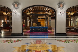 A grand lobby with a piano, a reception desk, and a decorative carpet with the text "Queen of the Gulf" and "Est. 1911".