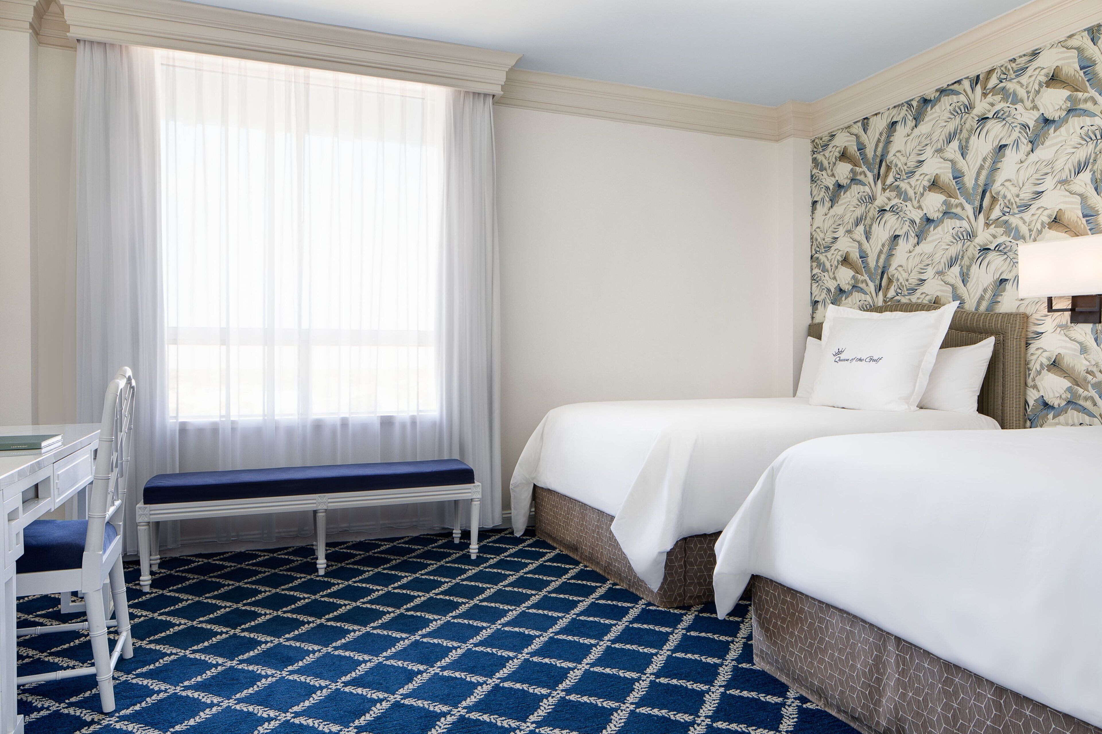 A hotel room with a large window, a desk, two beds, and a patterned floor.