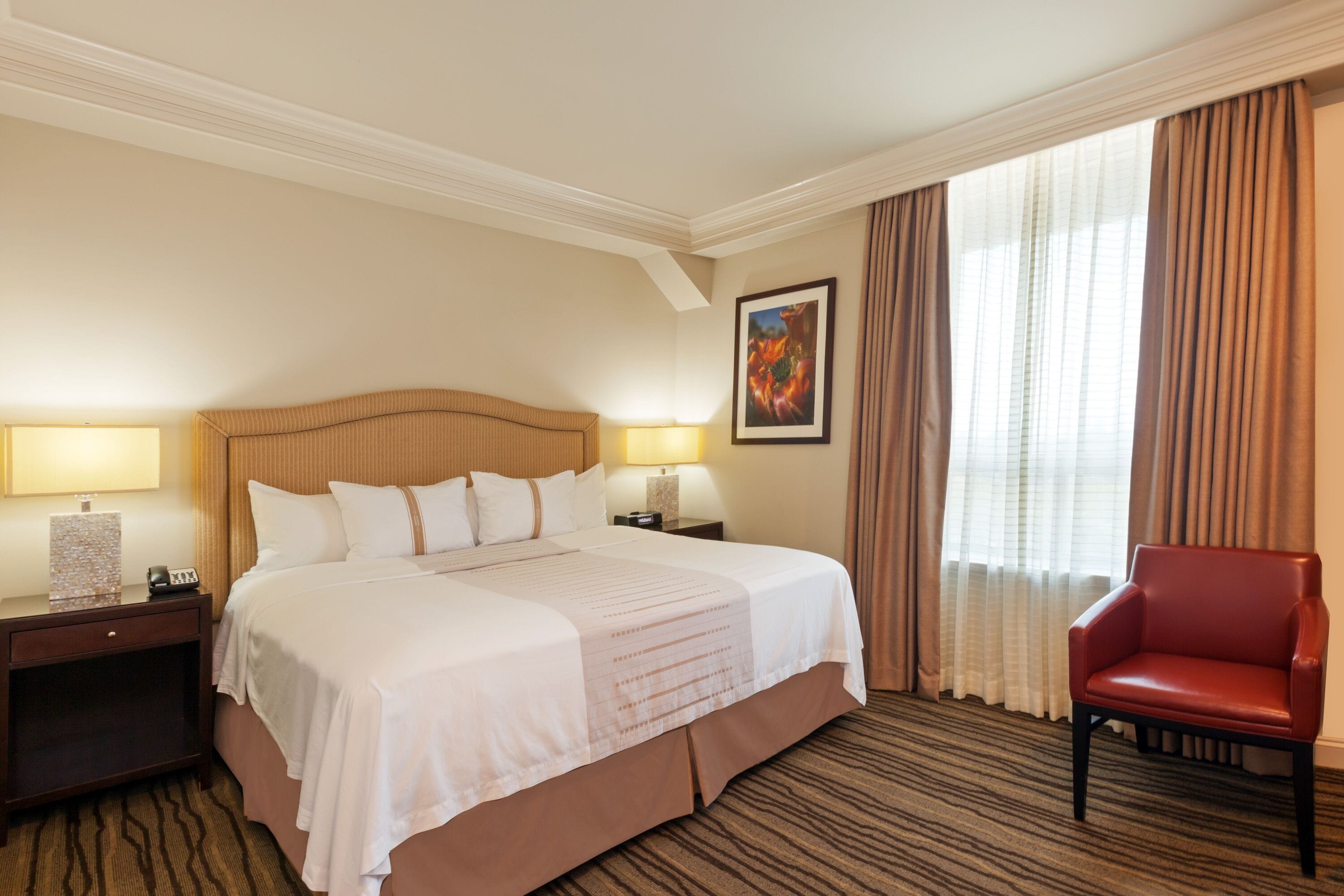 Une chambre d’hôtel avec un grand lit, deux tables de chevet avec des lampes, un fauteuil rouge et un tableau au mur.