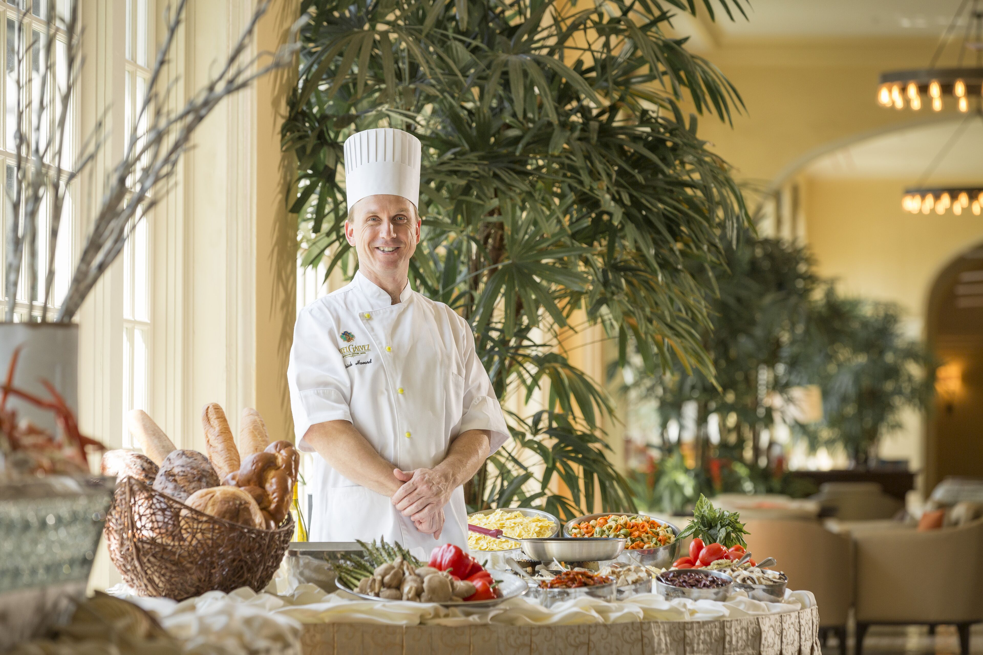 A chef standing in a well-lit room with a buffet table filled with various dishes and a large plant in the background.