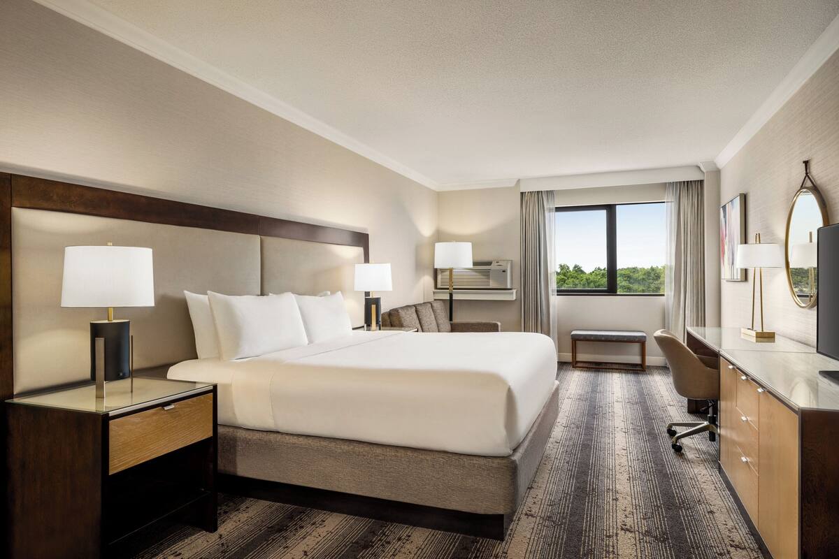 A hotel room with a large bed, bedside tables with lamps, a desk with a chair, and a view of the outdoors.