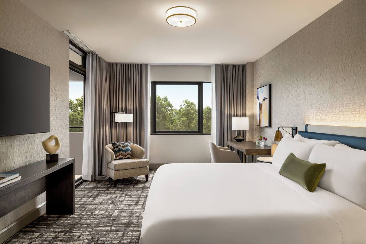 A hotel room with a large bed, a desk, a chair, a TV, and a window with curtains.