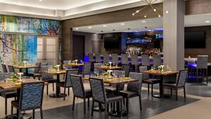 A modern restaurant with multiple dining tables and chairs, a bar area with a variety of bottles, and a large flat-screen TV mounted on the wall.