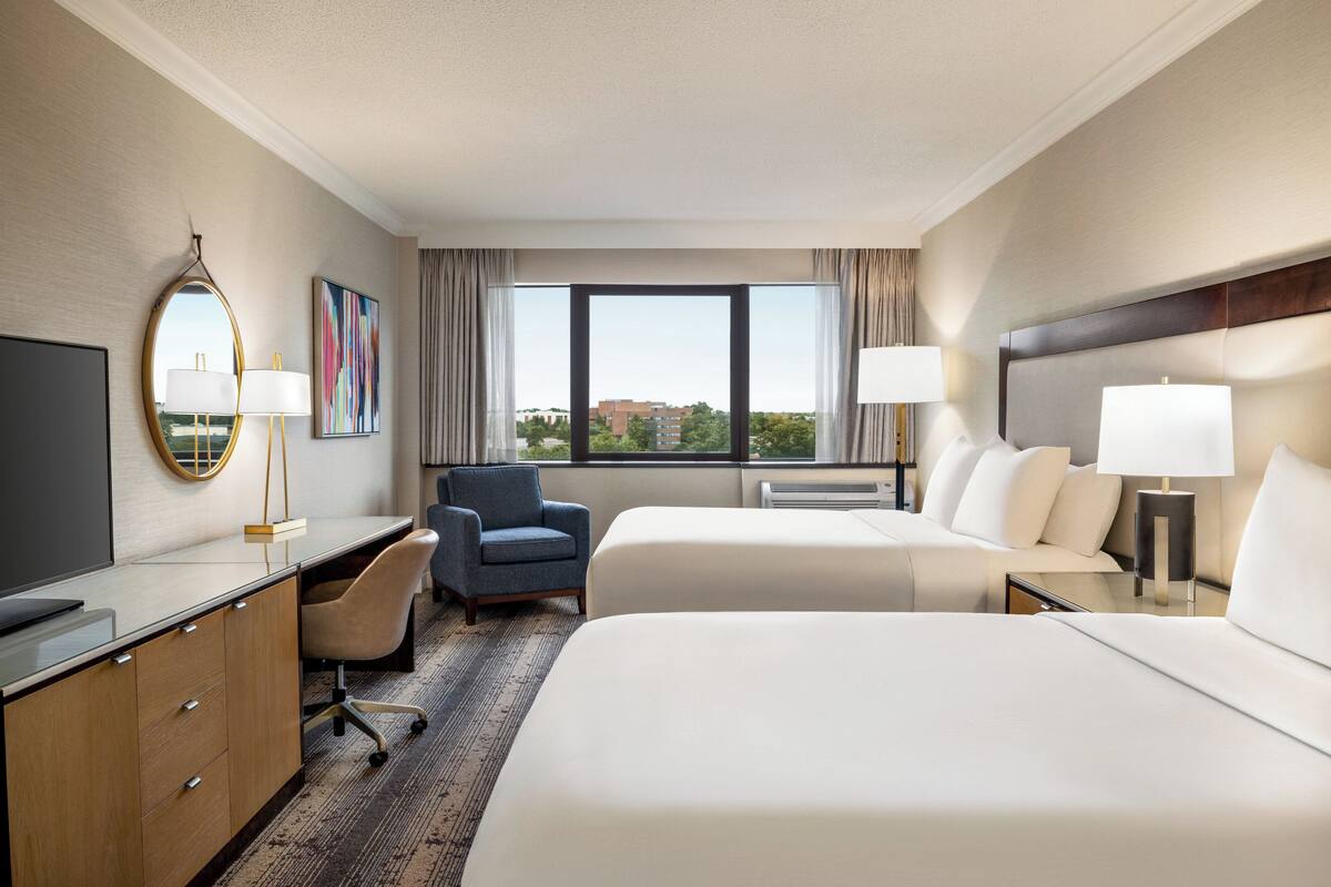 A hotel room with a desk, two beds, a chair, a large window, and a view of the city.