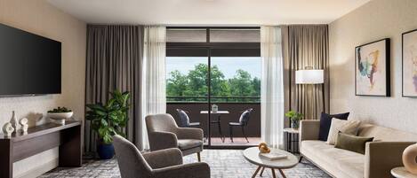 Habitación de hotel moderna con un televisor de pantalla plana grande, un sofá, dos sillones, una mesa de centro y un balcón con vistas a zonas verdes.
