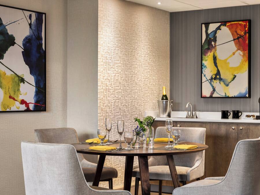 Un comedor moderno con una mesa redonda dispuesta para seis personas, rodeada de sillas tapizadas en gris. Hay dos cuadros abstractos en las paredes, un panel de pared con textura y una zona de cocina con un fregadero y una máquina de café.