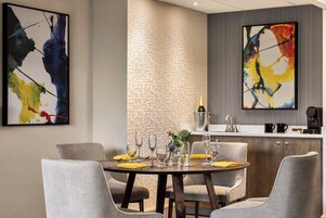 Un comedor moderno con una mesa redonda dispuesta para seis personas, rodeada de sillas tapizadas en gris. Hay dos cuadros abstractos en las paredes, un panel de pared con textura y una zona de cocina con un fregadero y una máquina de café.