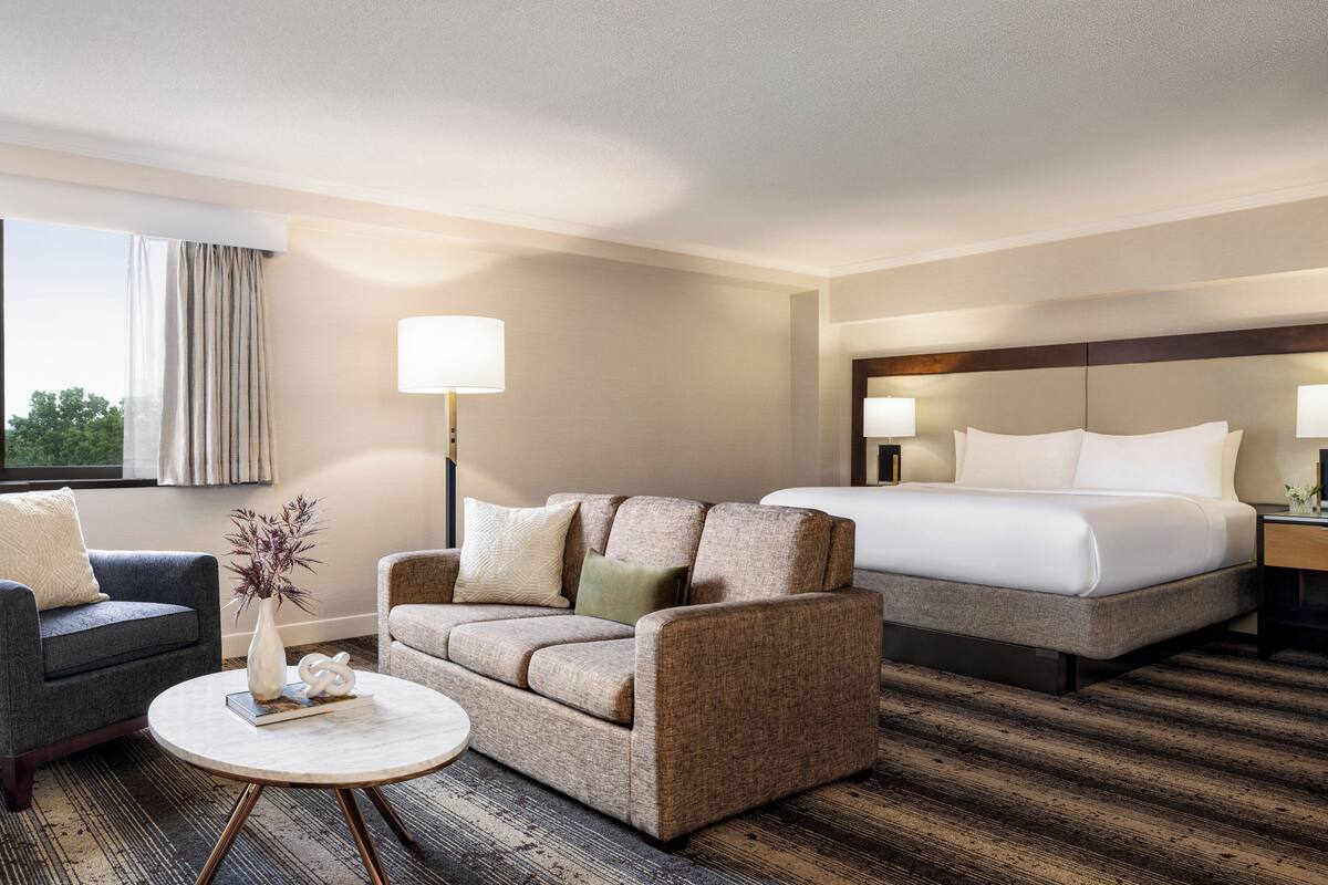 A hotel room with a bed, two armchairs, a round coffee table, and a lamp.