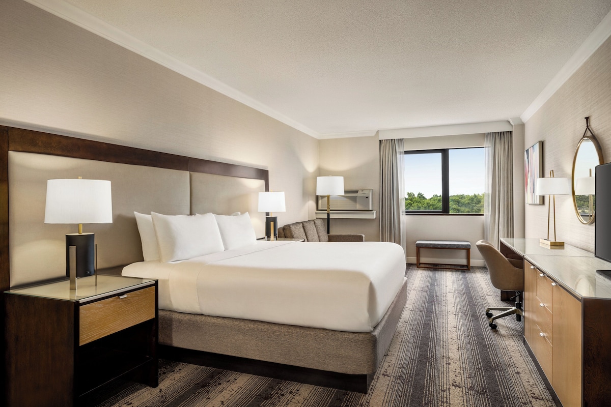 A hotel room with a large bed, bedside tables with lamps, a desk with a chair, and a view of the outdoors.