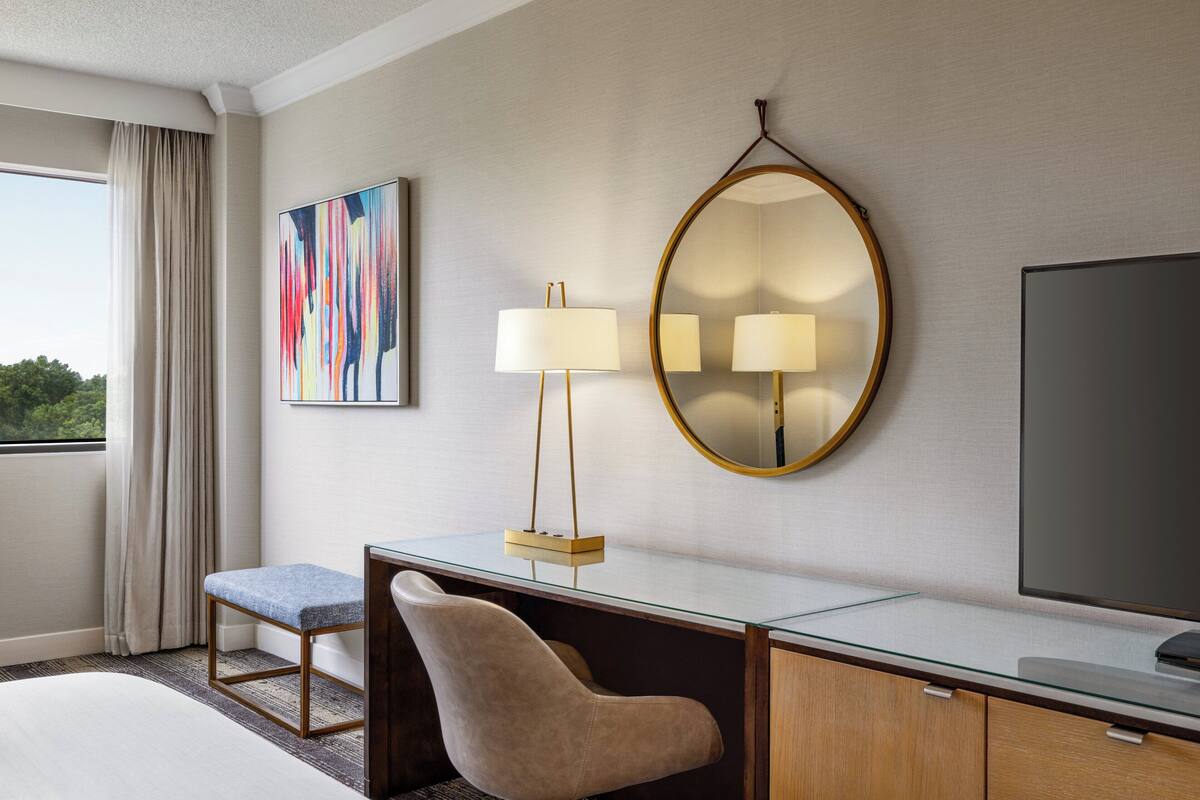 A hotel room with a bed, a desk with a chair, a large mirror, a television, and a window with curtains.