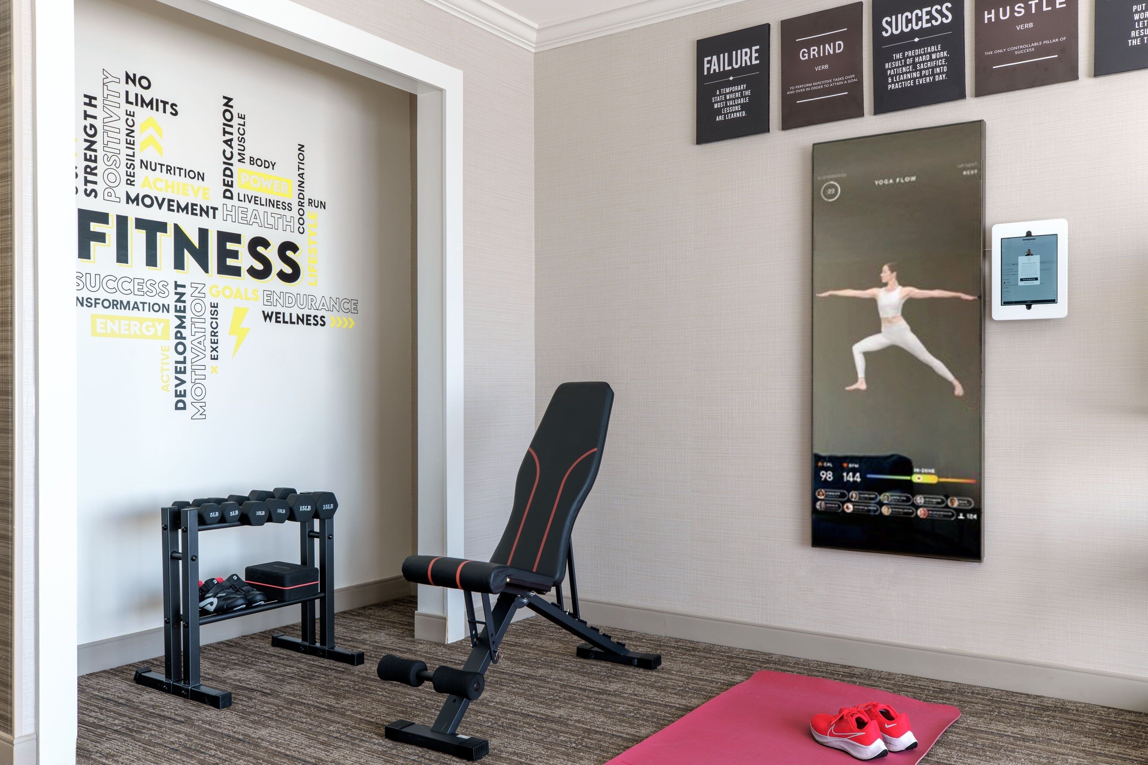 A fitness room with a wall-mounted poster, a standing desk, dumbbells, and a red mat.