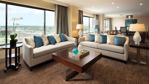 Habitación de hotel con dos sofás, una mesa de centro y una mesa de comedor con lámparas. Amplias ventanas ofrecen vista a la ciudad.
