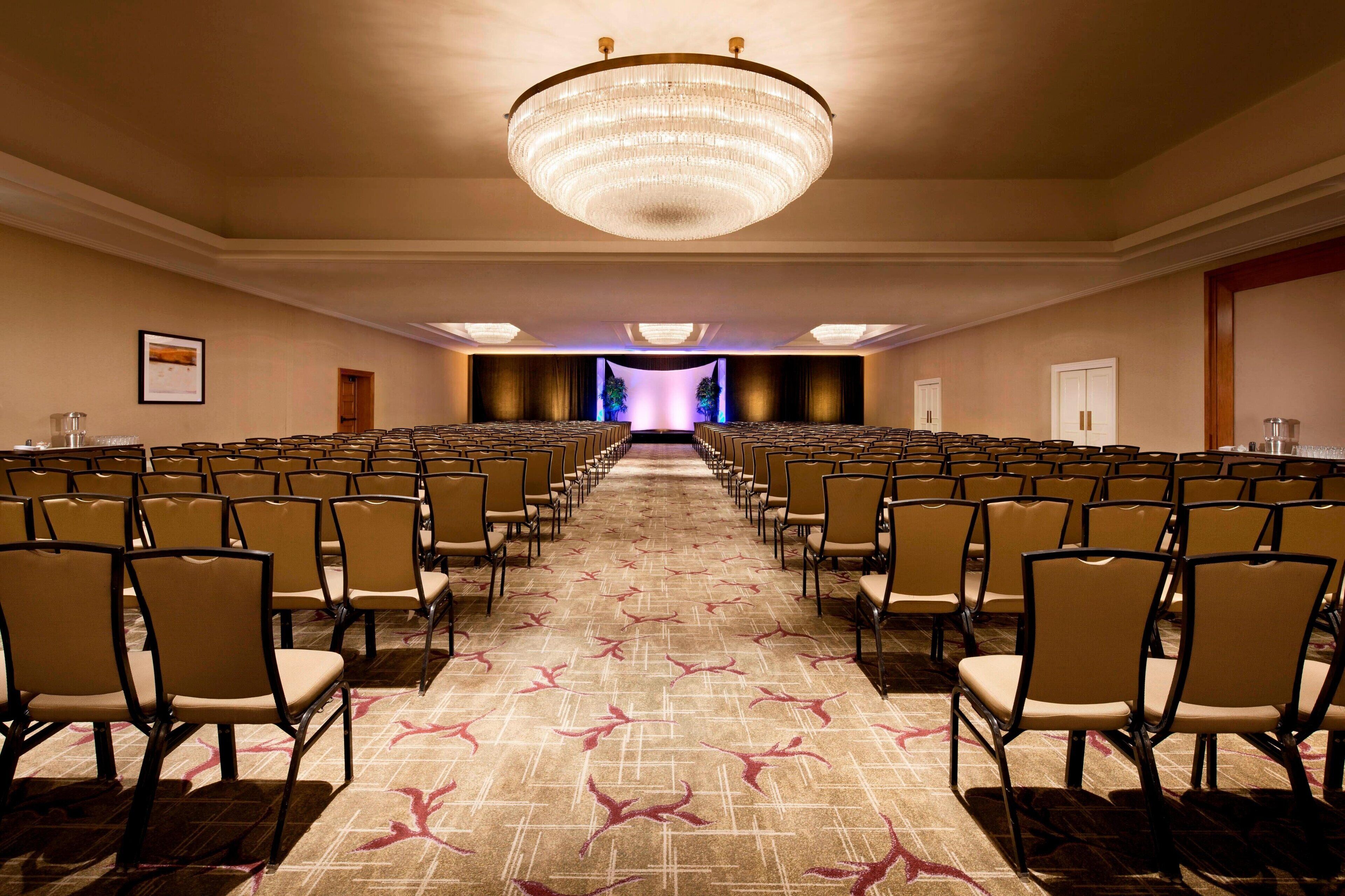 A conference room with rows of chairs, a large chandelier, and a carpeted floor.