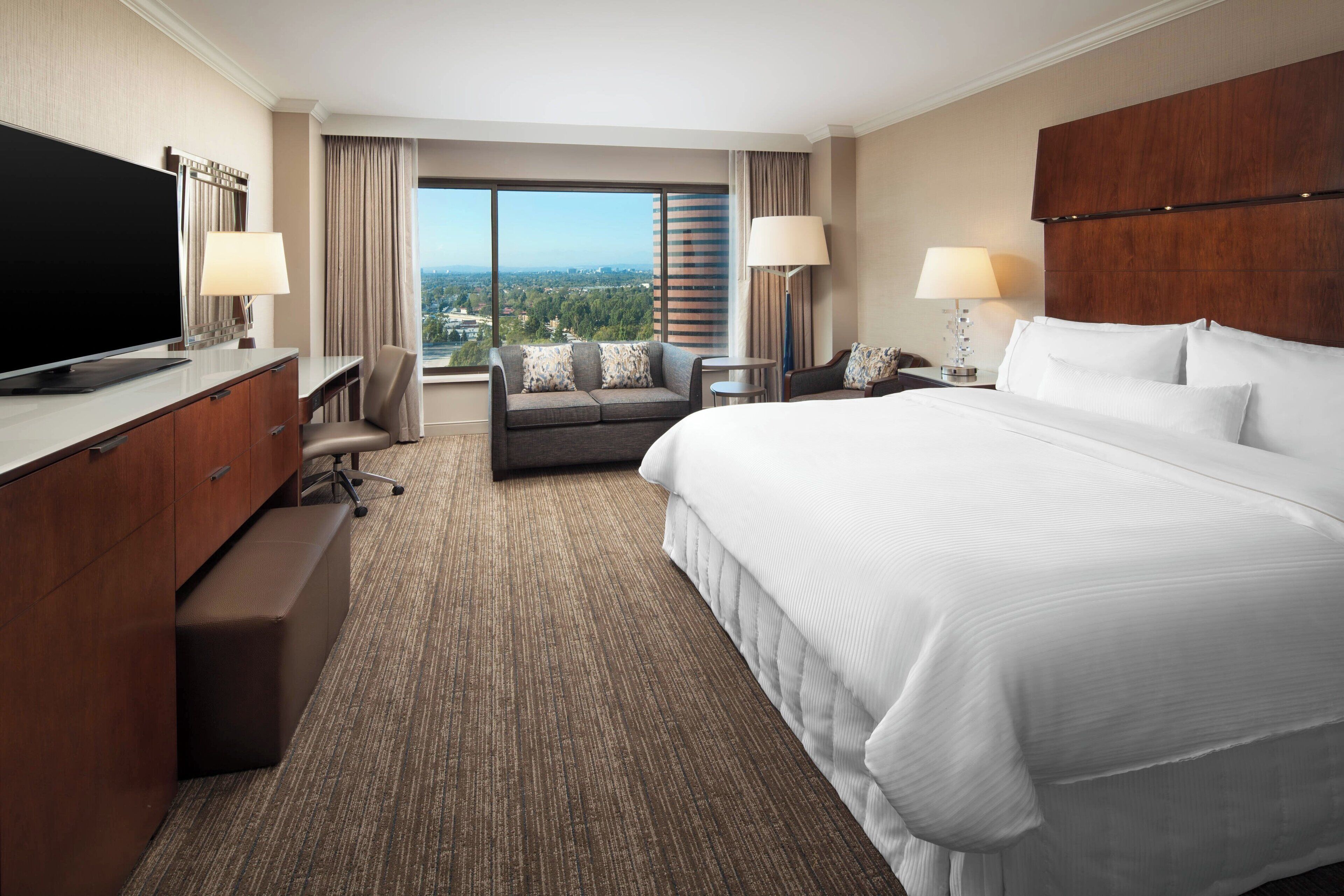 A hotel room with a large bed, a desk with a TV, a sofa, and a view of the city.