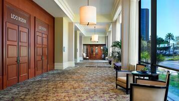 A hotel hallway with two LIDO ROOM doors, a patterned carpet, a chandelier, seating area with a table, and a view of the street.