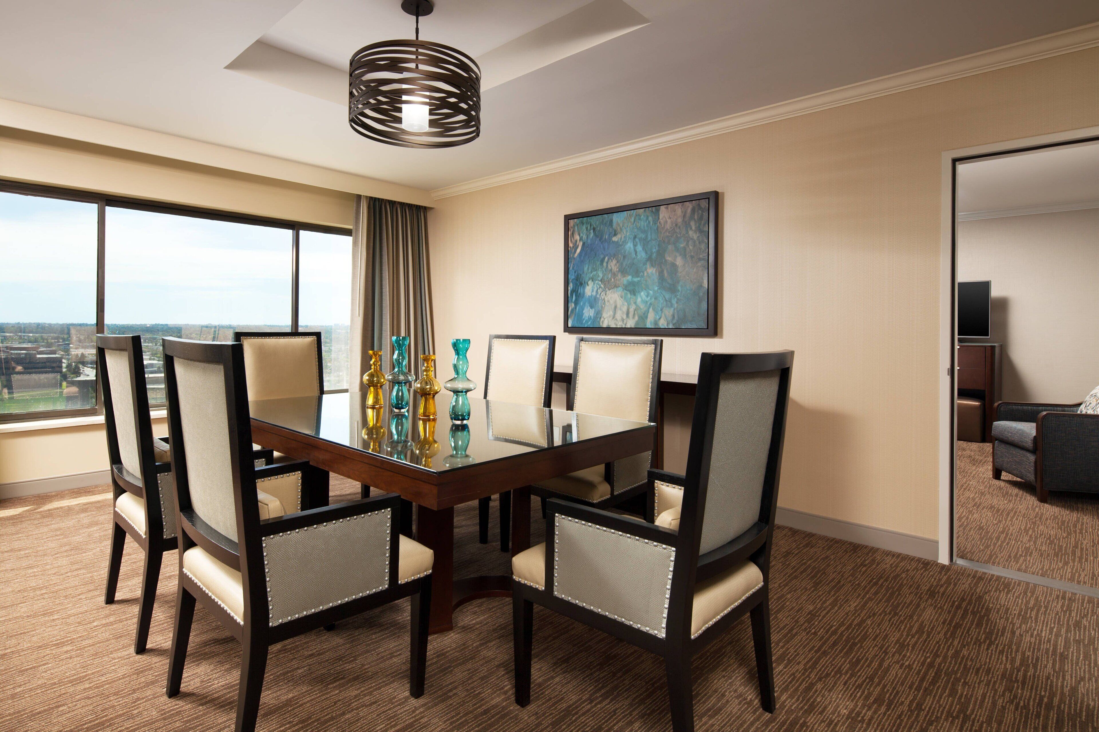 A hotel dining room with a large table, six chairs, and a city view through large windows.