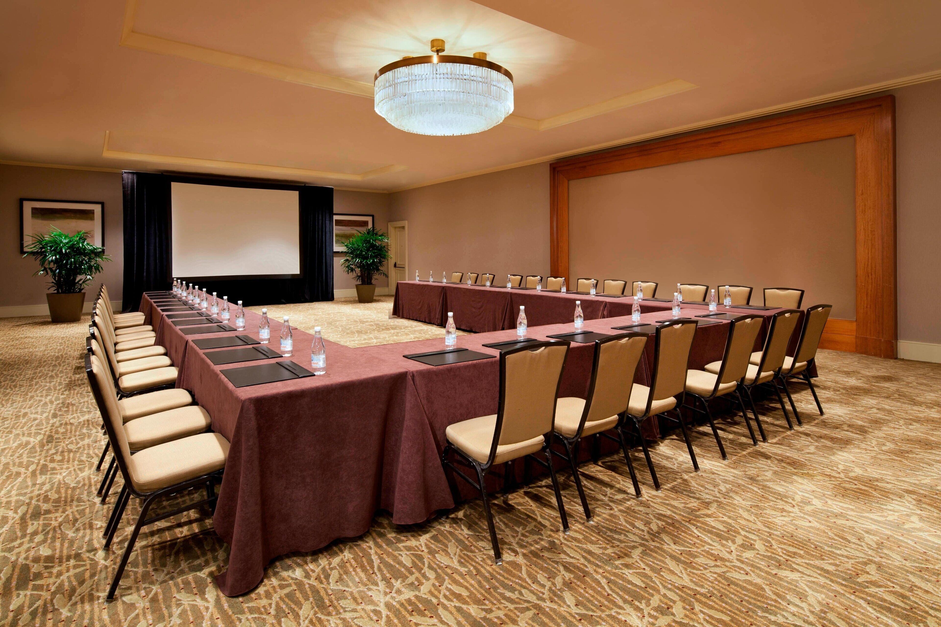 A conference room with a long table, chairs, a projector screen, and a chandelier.
