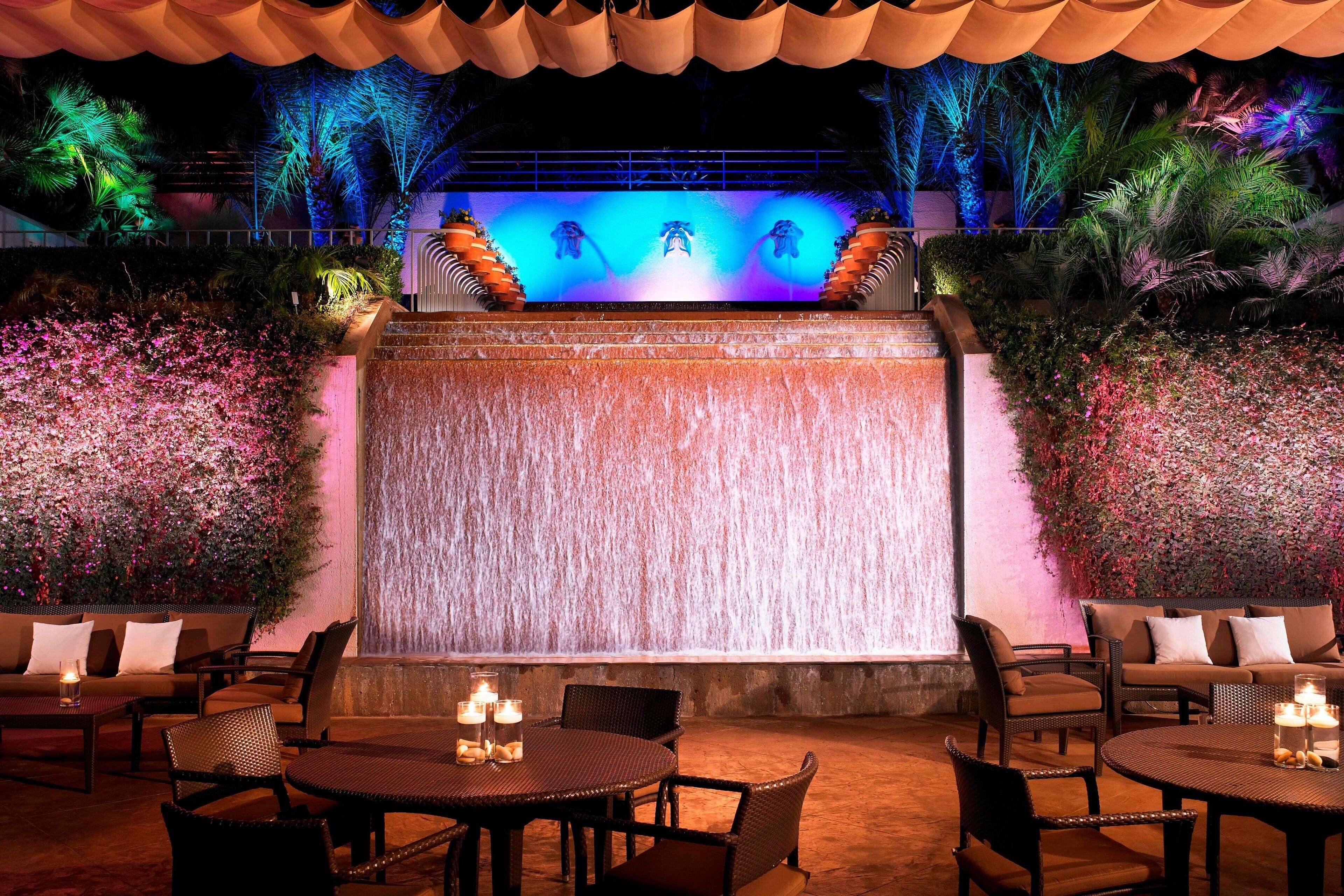 An outdoor seating area with tables and chairs, a waterfall feature, and a backdrop of foliage and palm trees.