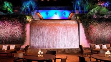 An outdoor seating area with tables and chairs, a waterfall feature, and a backdrop of foliage and palm trees.