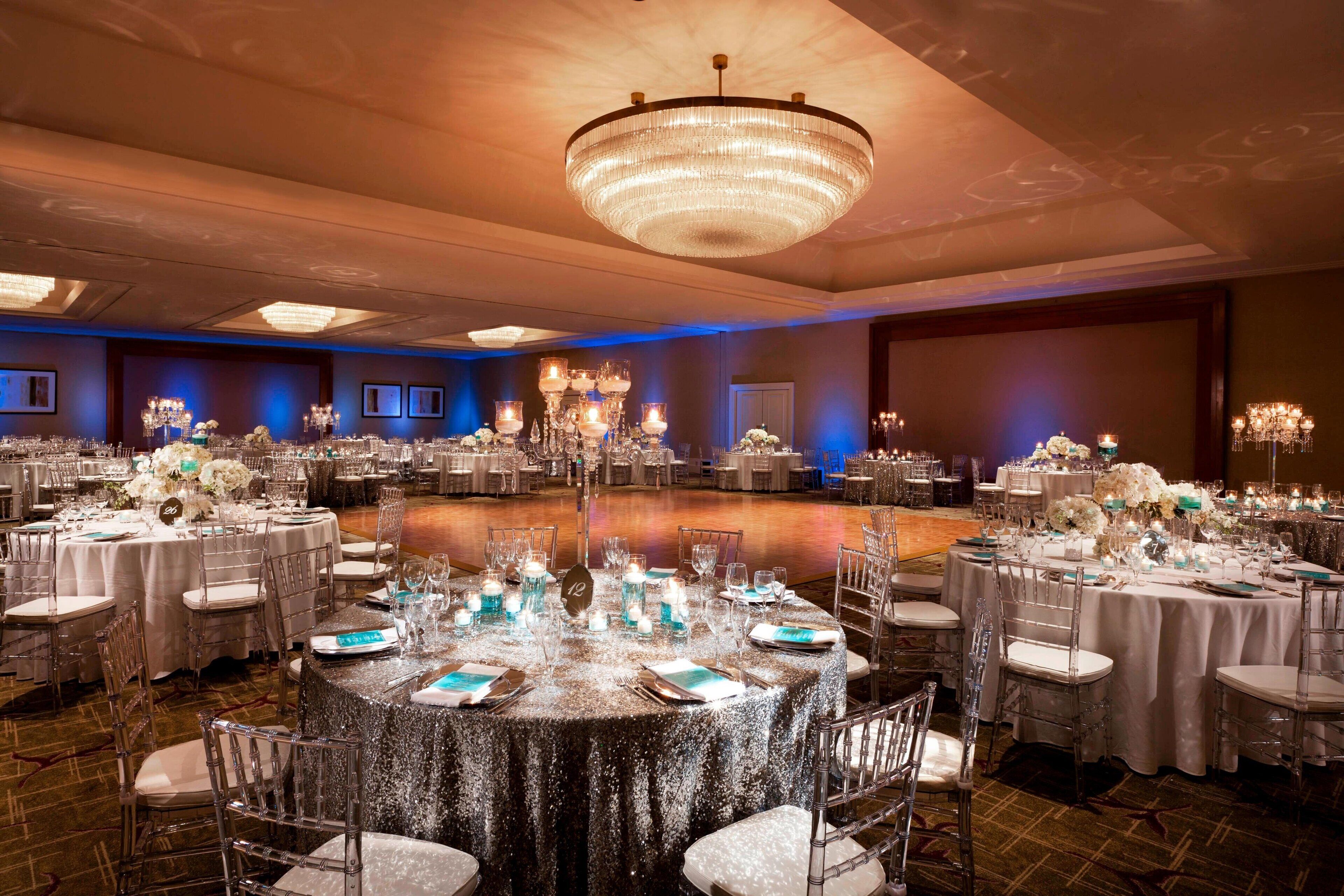 A banquet hall with round tables, clear chairs, and chandeliers.
