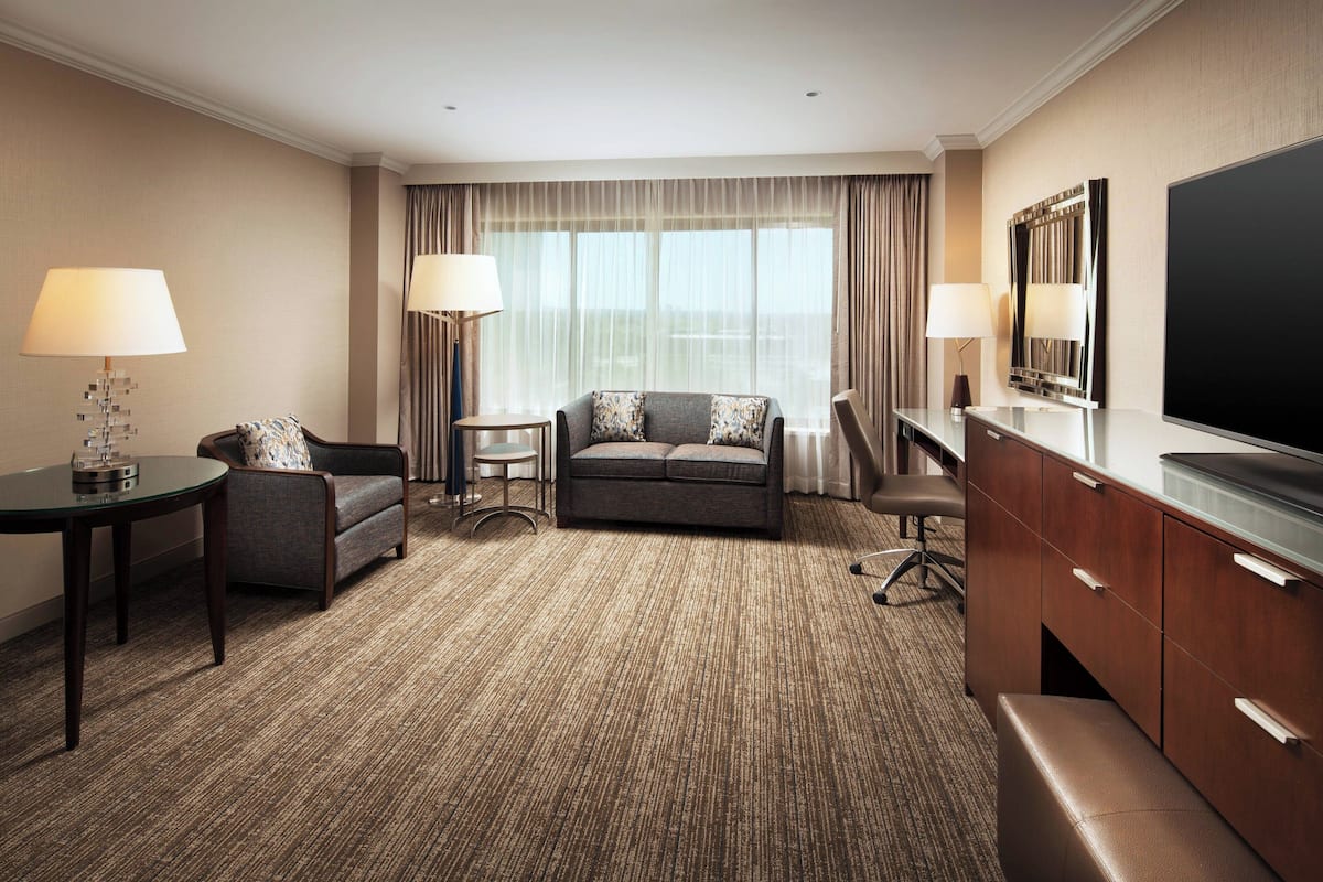 Una habitación de hotel con sofá, sillón, una mesita con lámpara, televisor y un gran ventanal con cortinas.