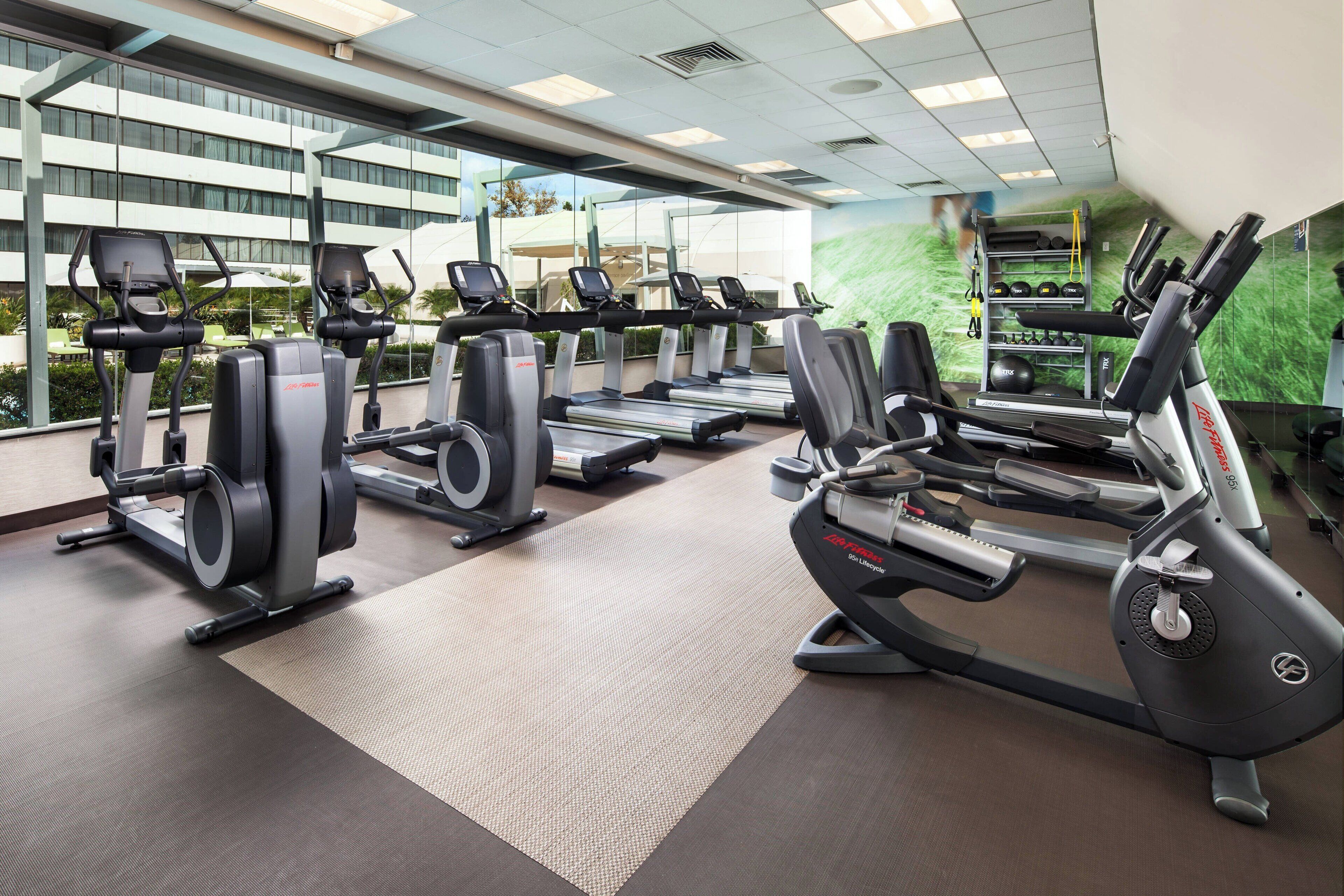 Ein Fitnessstudio mit Laufbändern, Crosstrainern und Gewichtsmaschinen, großen Fenstern und Blick auf ein Gebäude.