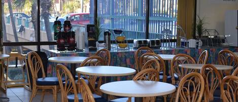 Un restaurante con sillas de mimbre y mesas redondas, vista a palmeras y una cerca azul en el exterior, y un letrero que dice 'Daybreak Breakfast'.