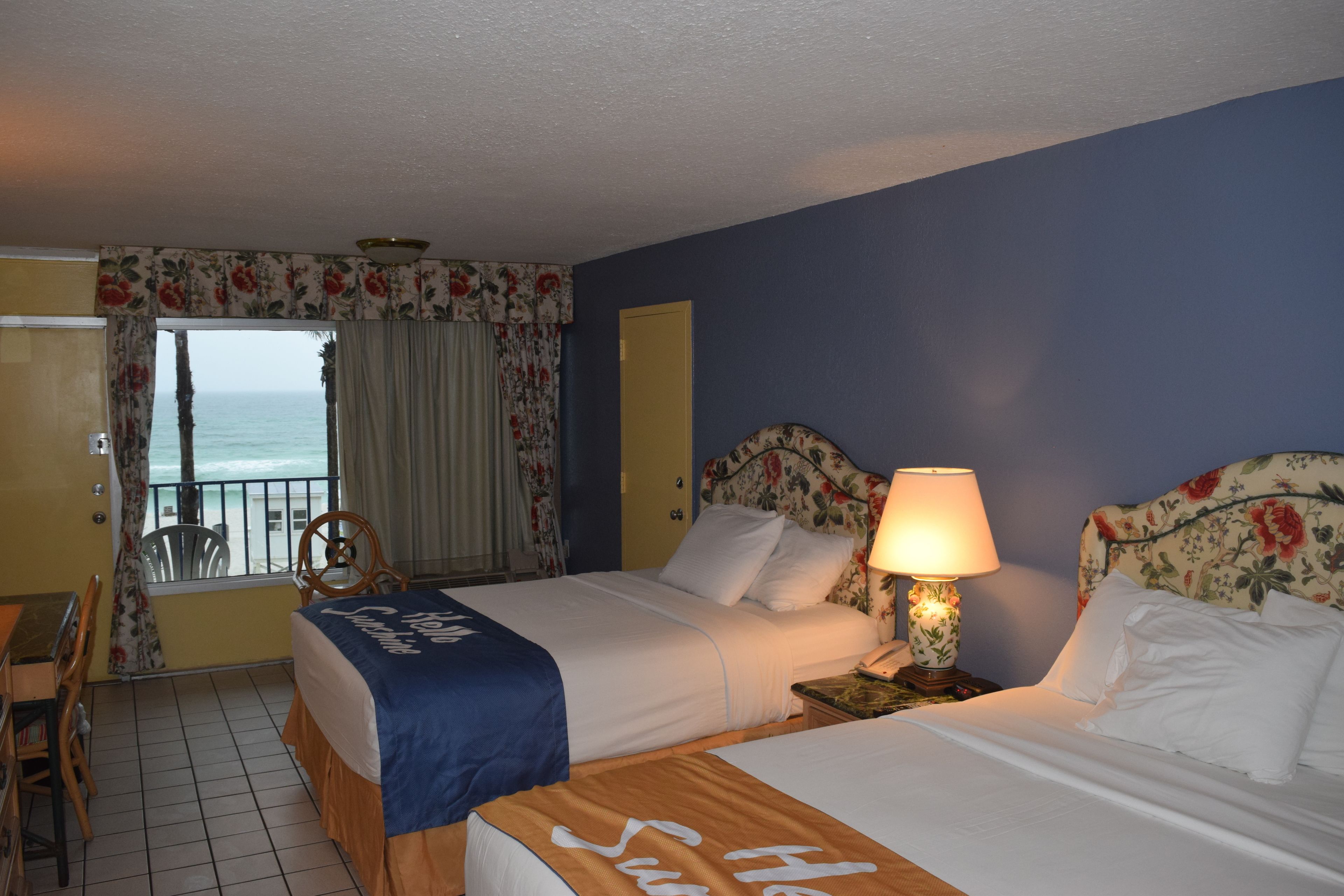 A hotel room with two beds, a balcony with a view of the sea, a desk with a chair, and a small table with a lamp.