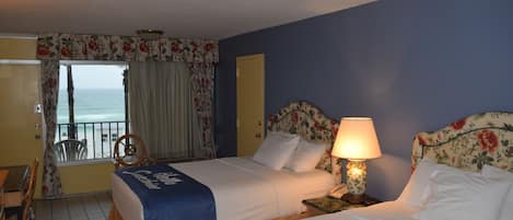Ein Hotelzimmer mit zwei Betten, einem Balkon mit Meerblick, einem Schreibtisch mit Stuhl und einem kleinen Tisch mit Lampe.