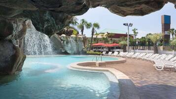 A pool with a waterfall feature, surrounded by lounge chairs and umbrellas, with a building in the background.