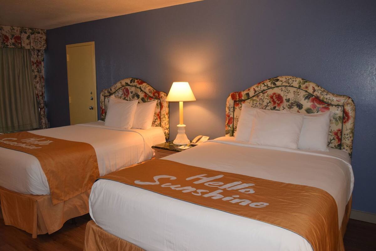Une chambre d’hôtel avec deux lits, des têtes de lit à motifs floraux, une table de chevet avec une lampe et une pancarte « Bonjour, soleil ! » sur les lits.