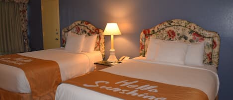 Habitación de hotel con dos camas, cada una con cabecera adornada con motivos florales y una colcha con el estampado 'Hello Sunshine'. Entre las camas, hay una mesita de noche con una lámpara y un teléfono.