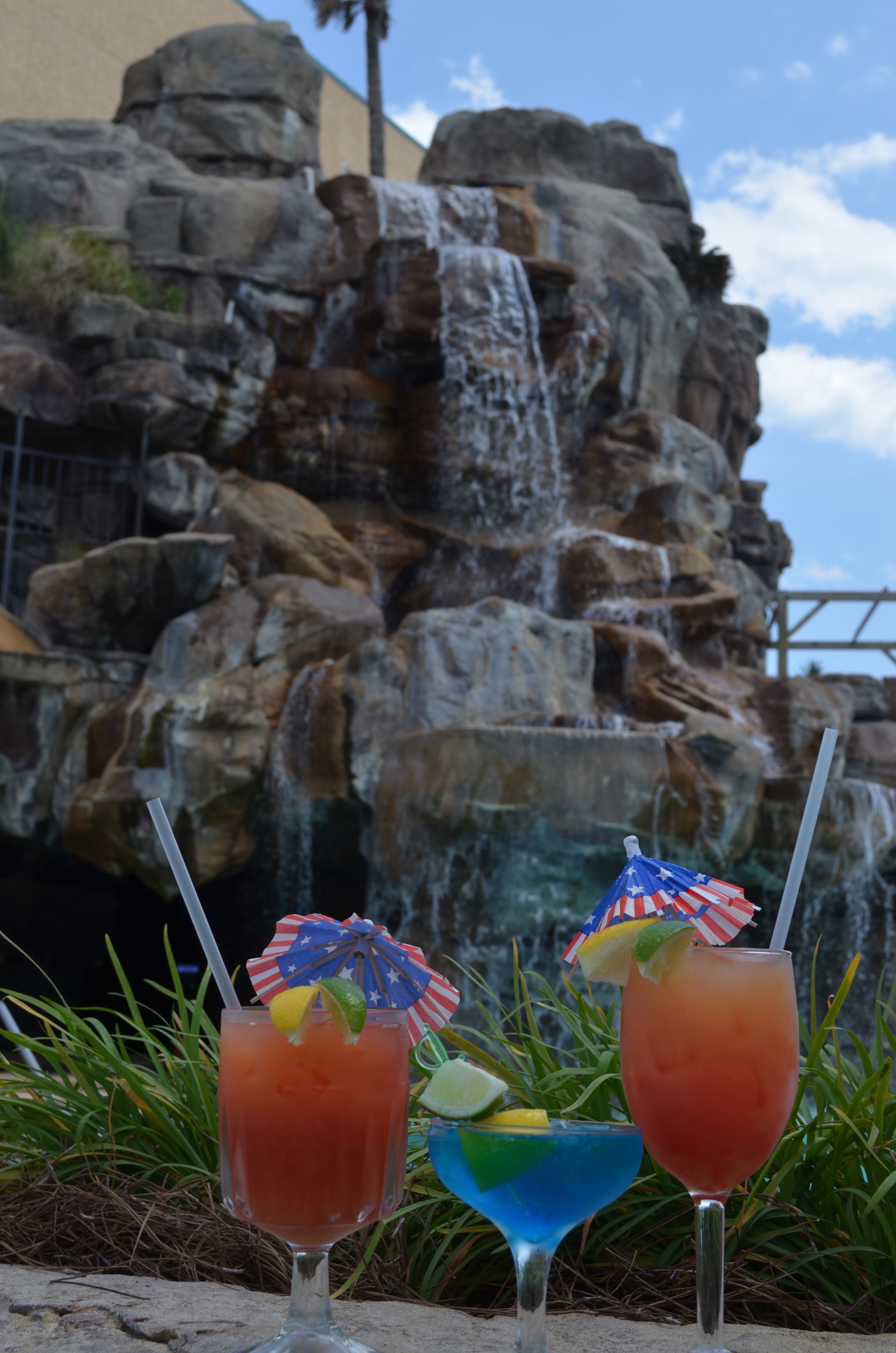 Two cocktails with umbrellas and straws in front of a rocky waterfall.