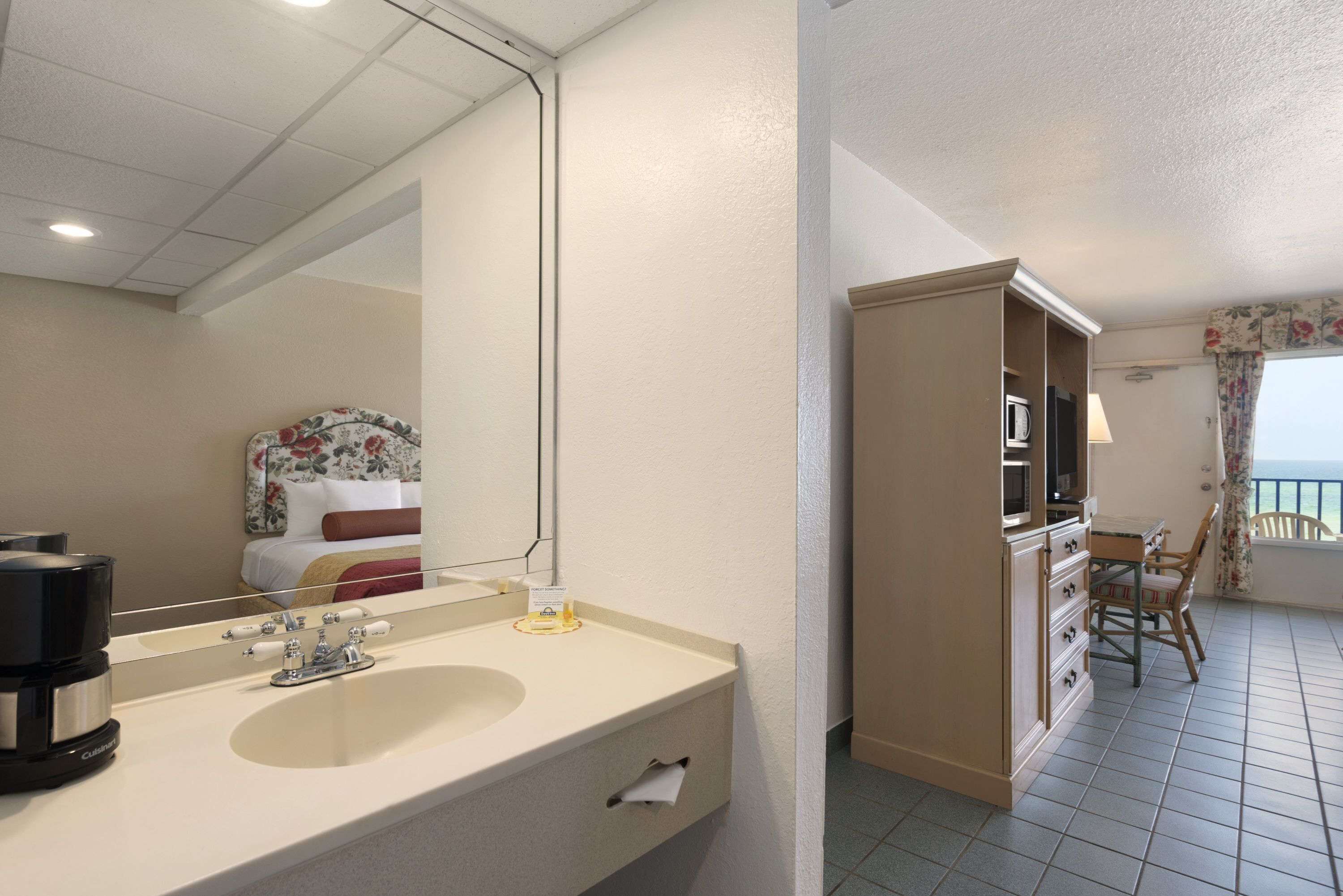 A hotel room with a large mirror, a double sink, a coffee maker, a TV, and a balcony with a chair.