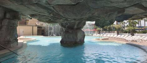 Zona de piscina con una gran formación rocosa sobre ella, agua cristalina y sillones de descanso alrededor del borde.