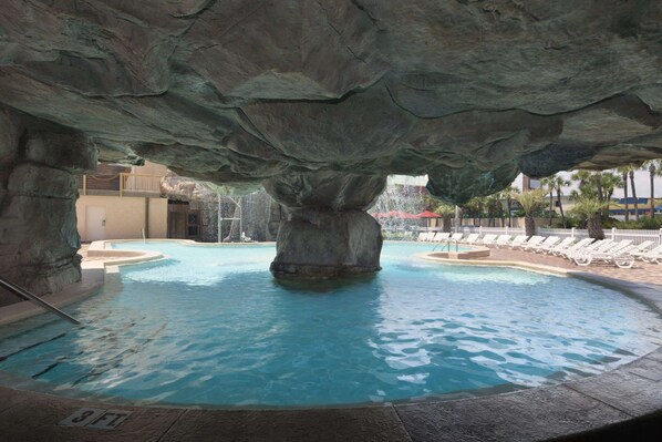 Un espace piscine agrémenté d’une grande formation rocheuse au-dessus, d’une eau cristalline et de chaises longues disposées tout autour.