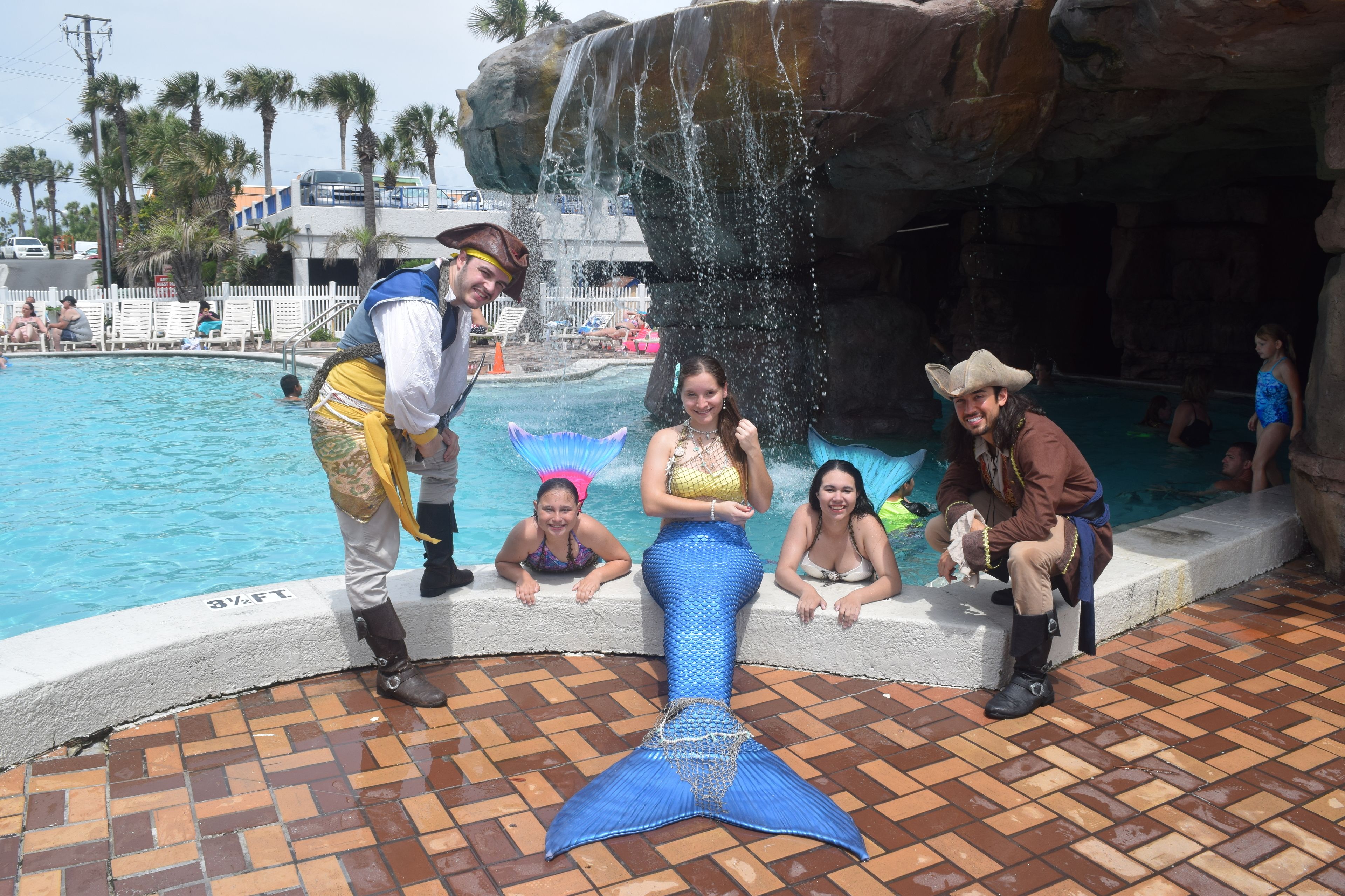 A pool area with a mermaid tail decoration, palm trees, and a rocky waterfall backdrop.