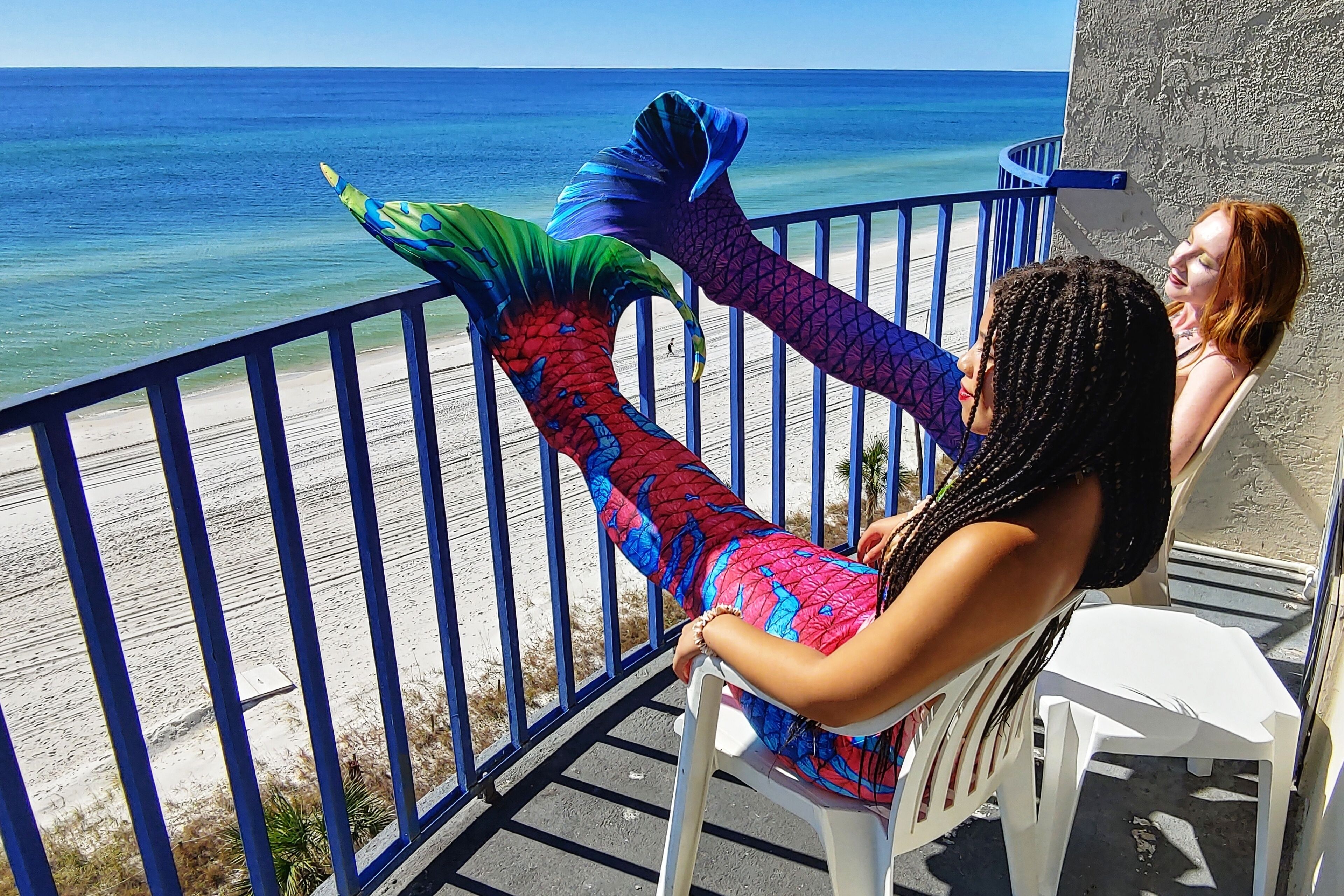 Un balcone con vista sull'oceano, una persona travestita da sirena e una sedia di plastica bianca.