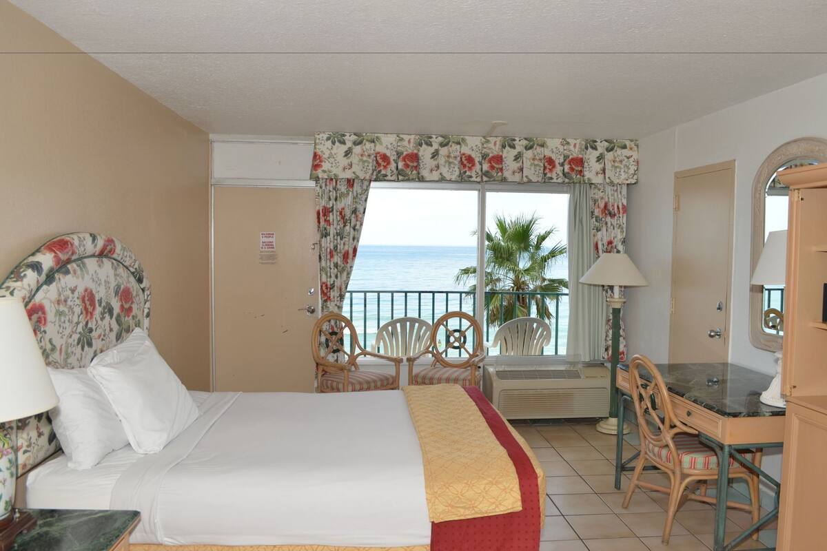 Une chambre d’hôtel avec un lit, un bureau, une chaise, une fenêtre donnant sur la mer et un balcon agrémenté d’un palmier.