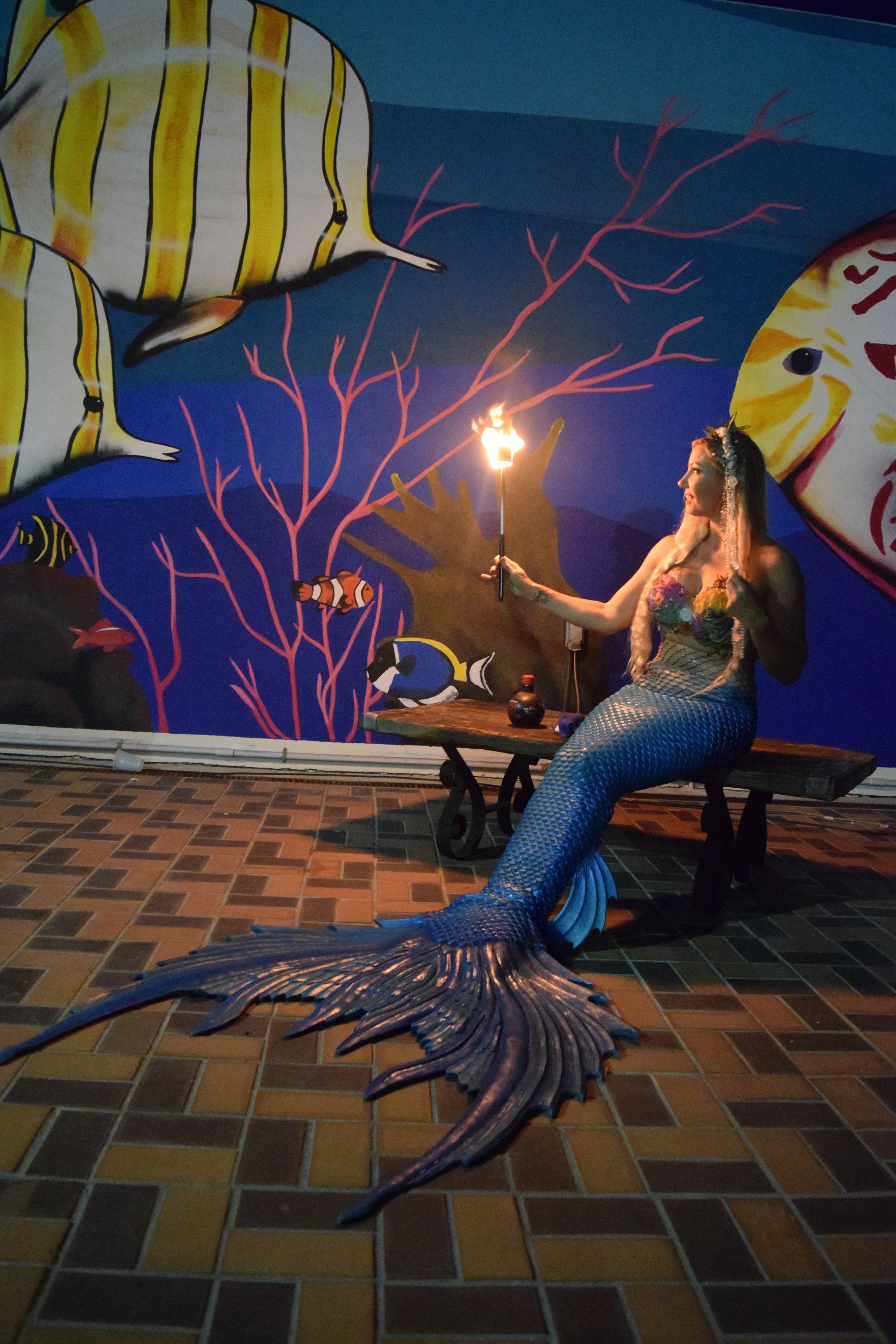 A mermaid statue with a torch, seated on a bench with a mural of underwater scenery in the background.
