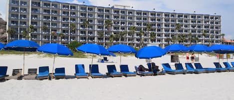 Ein Hotel mit mehreren Balkonen, blauen Sonnenschirmen und Liegestühlen am Strand.