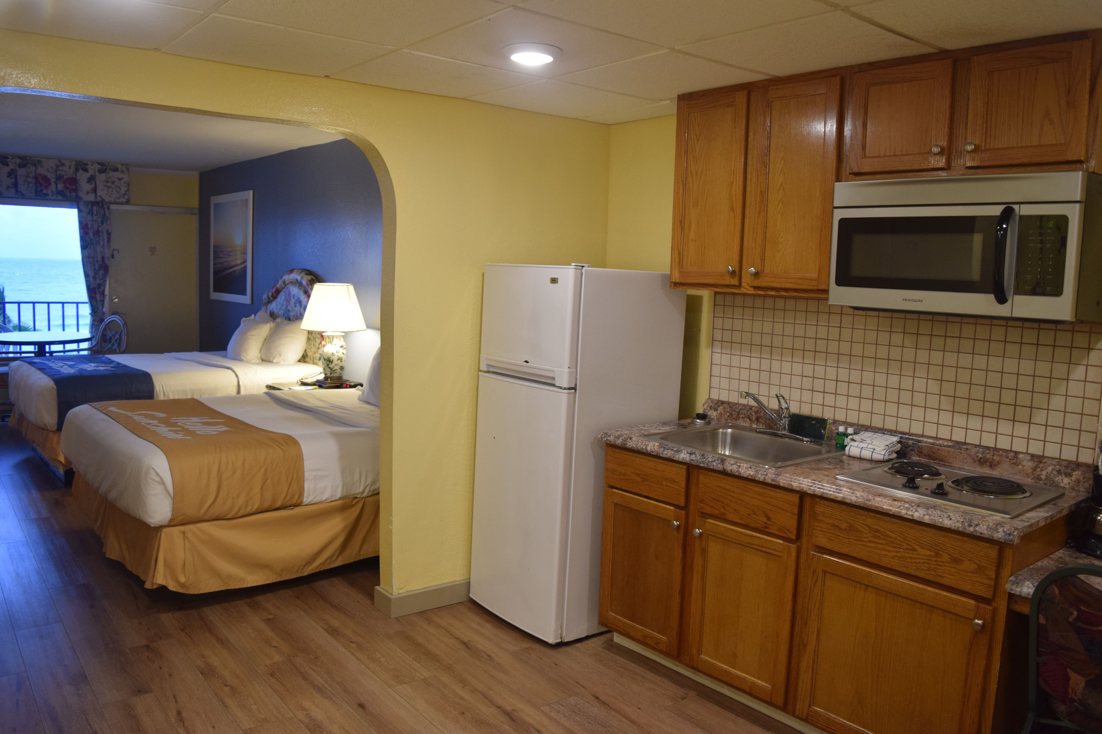 A compact hotel room with a bed, a small kitchenette, wooden cabinets, and a view of the ocean.