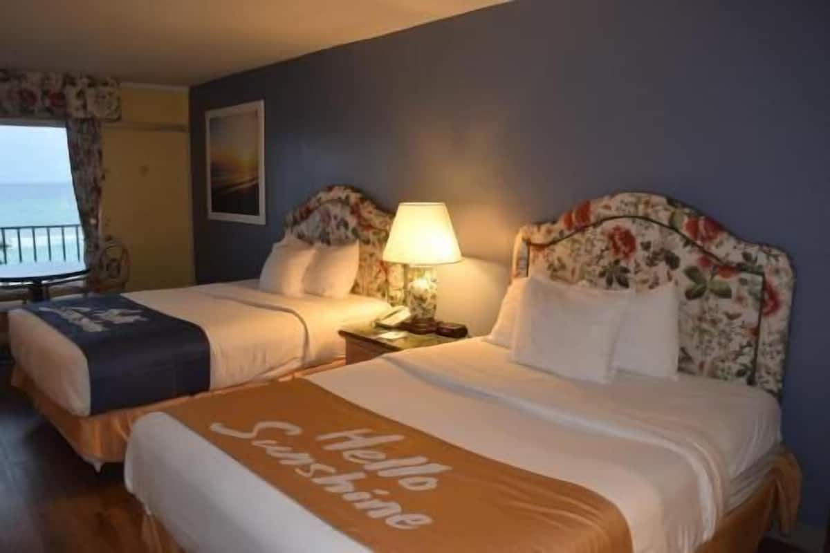 Una habitación de hotel con dos camas, una mesita de noche con lámpara, un cuadro en la pared y una ventana con cortinas.