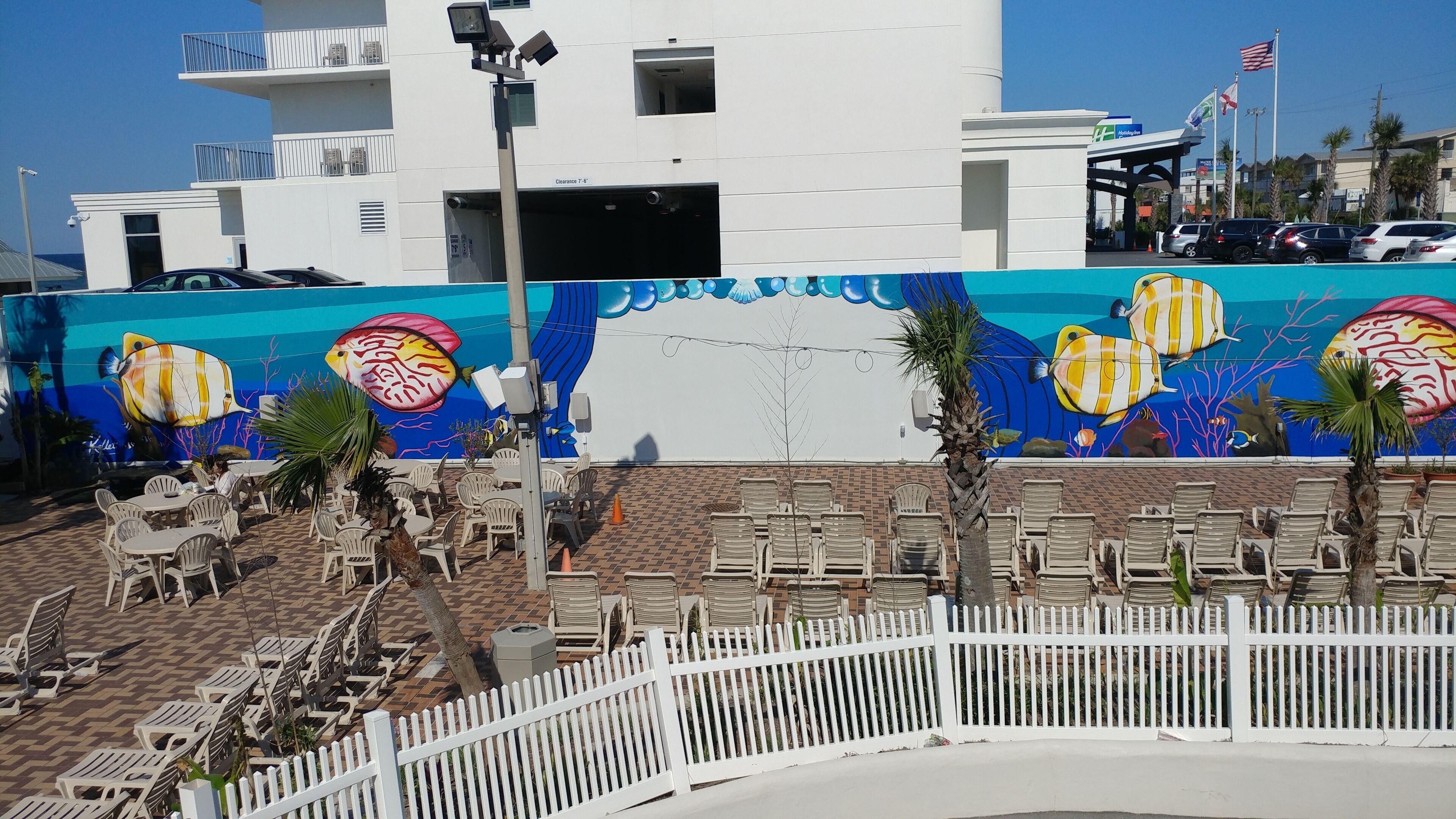 A hotel with a mural of tropical fish on the exterior wall, outdoor seating with white chairs, and a white picket fence.