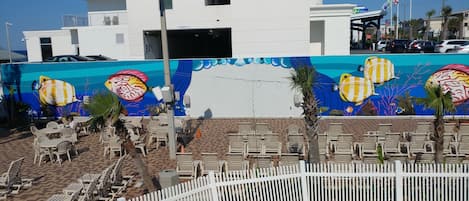 A hotel with a mural of tropical fish on the exterior wall, outdoor seating with white chairs, and a white picket fence.