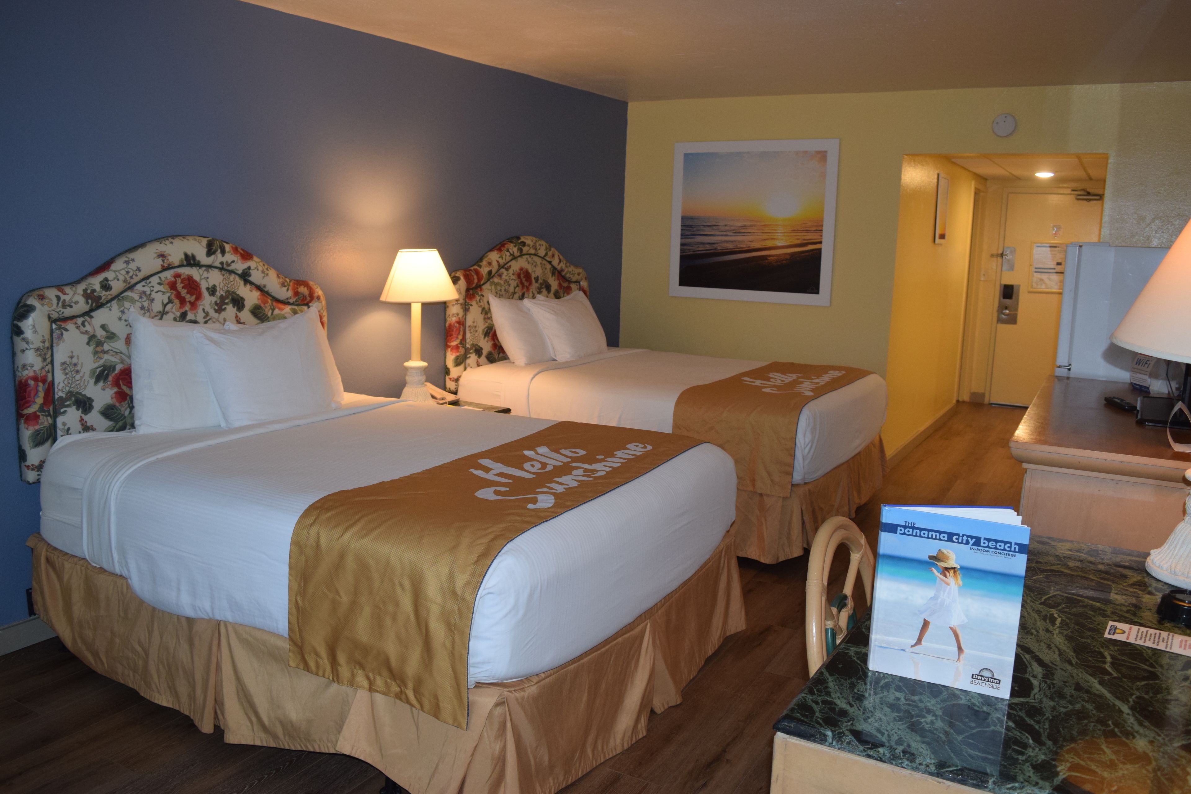 A hotel room with two beds, each with floral headboards and beige bedspreads. There is a nightstand with a lamp, a small table with a magazine, and a view of the hallway.