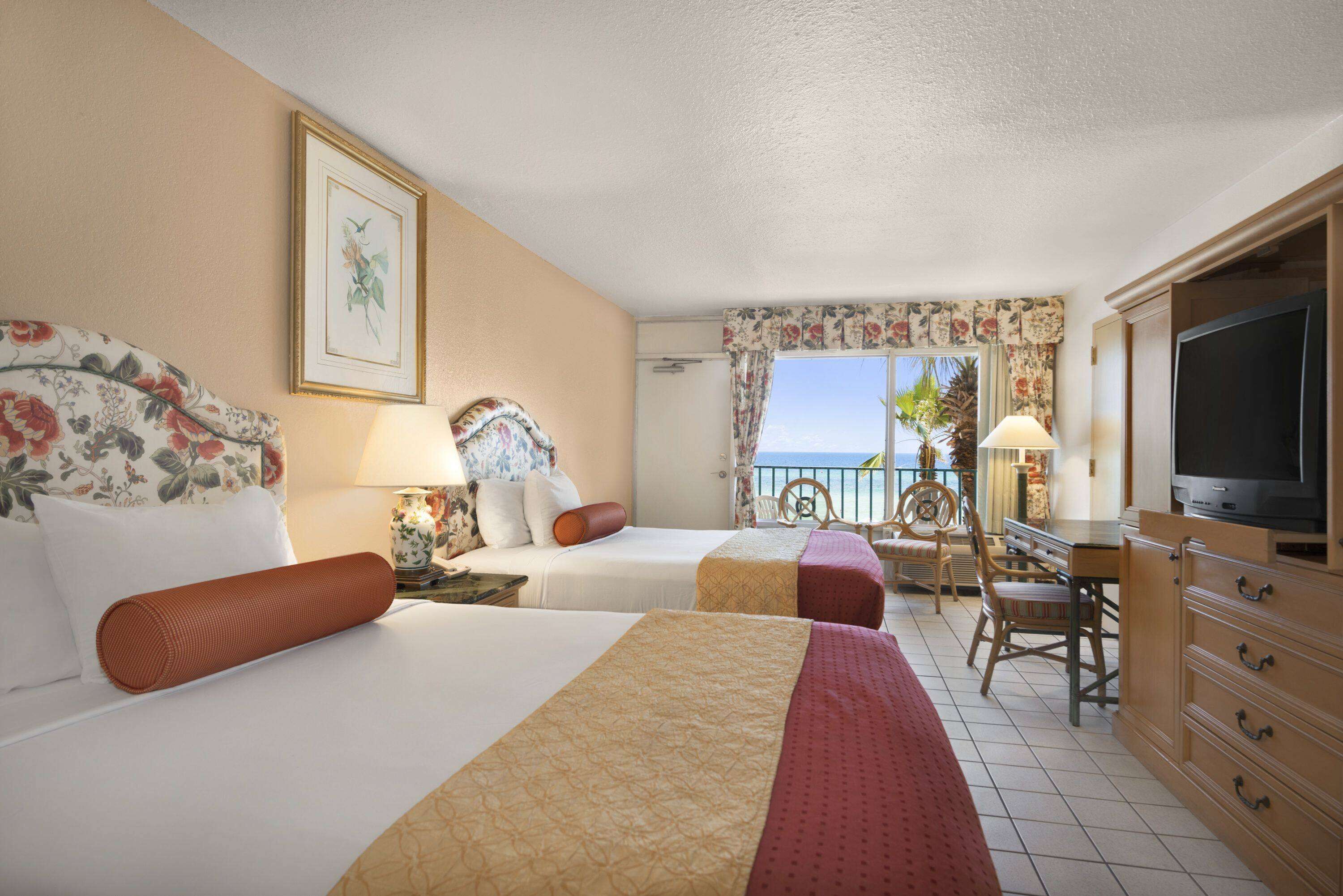 A hotel room with two beds, a television, a desk, and a view of the sea.