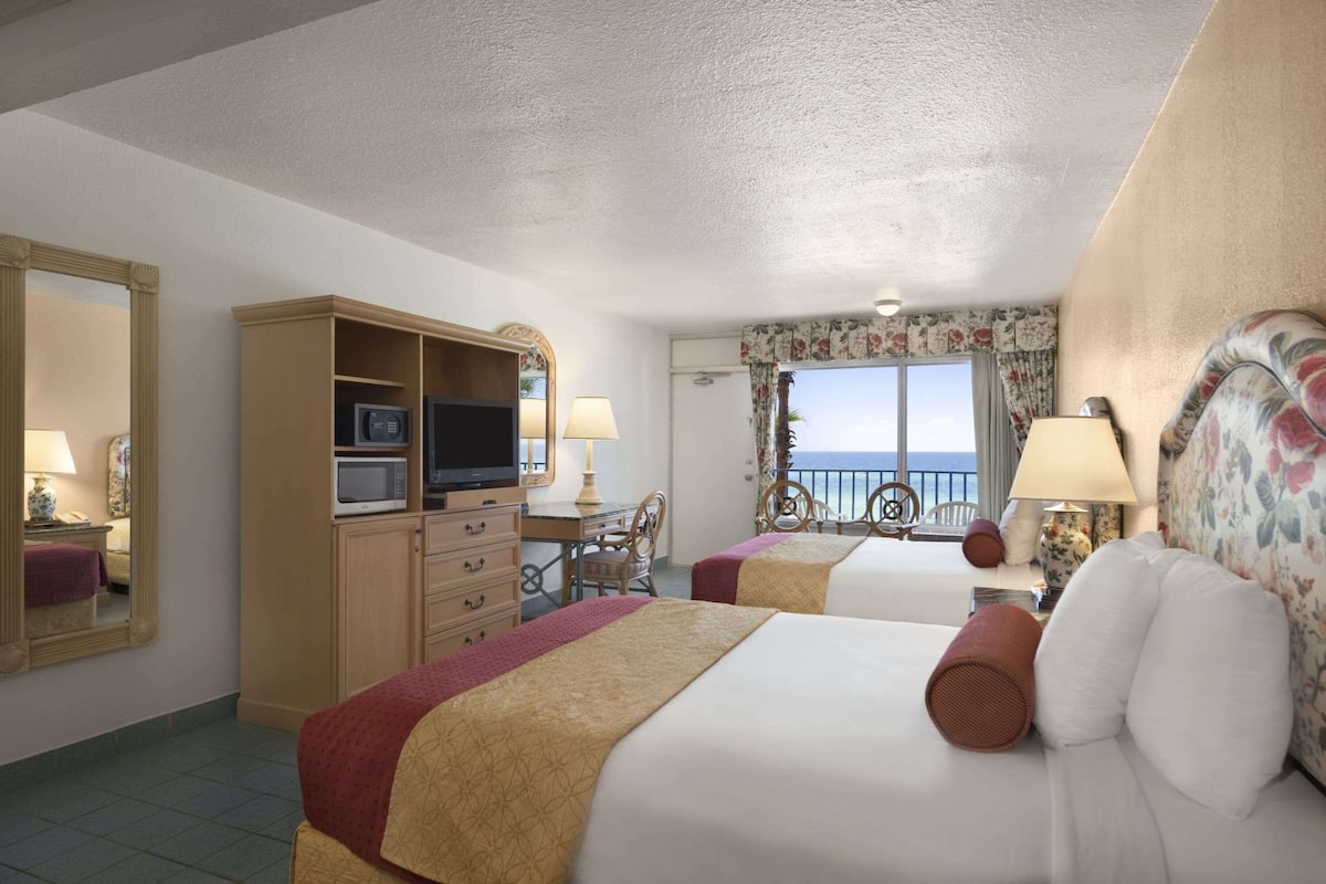 Habitación de hotel con dos camas, un escritorio con televisor, cómoda y vista al mar.